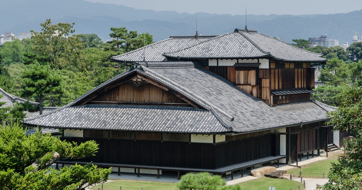 The Honmaru Palace at Nijo Castle, a two-storey timber palace among pines, seen from the keep tower base with Kyoto's hills behind
